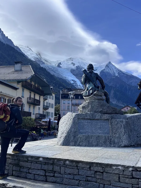 Eine Statue auf einem Sockel auf einem Platz in Chamonix, dahinter erheben sich direkt über den Dächern die frisch verschneiten Gipfel des Mont-Blanc-Massivs.