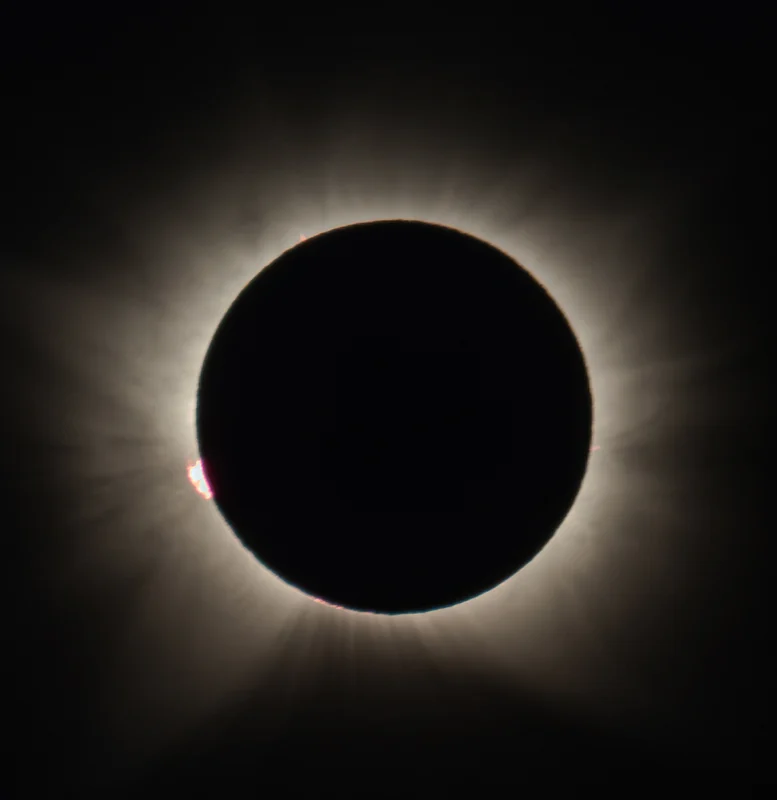 Die total verfinsterte Sonne: eine schwarze Mondscheibe, umgeben von der strukturierten weißen Korona, mit einer kleinen rosa Protuberanz am unteren linken Rand.
