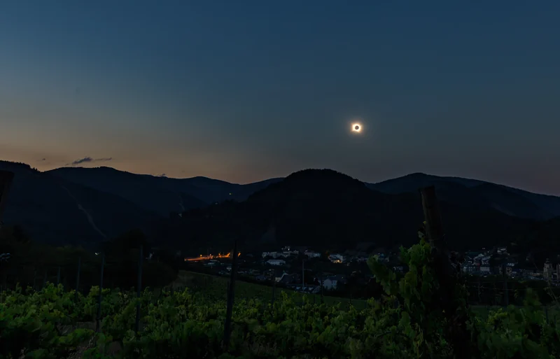 Die verfinsterte Sonne und ihre Korona in einem tiefblauen Dämmerungshimmel über einem spanischen Tal, im Vordergrund Rebzeilen, unten ein beleuchtetes Dorf.