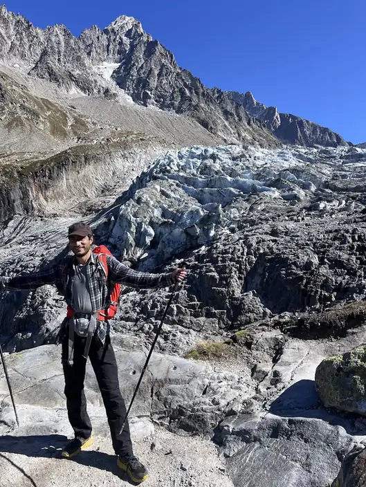 Eine Person mit Stöcken und Rucksack steht auf blankem Fels am Fuß eines Gletschers im Mont-Blanc-Massiv, dahinter aufgebrochene Séracs.