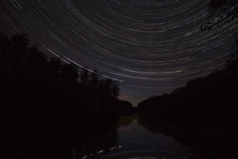 Zirkumpolare Strichspuren über stillem Wasser zwischen dunklen bewaldeten Ufern, die Bögen spiegeln sich im Wasser.